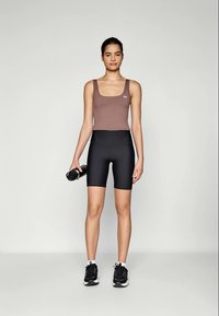 Smeđi atletski crop top, crne biciklističke kratke hlače visokog struka i crne tenisice. Model drži crnu bocu za vodu, pozira protiv neutralne pozadine.