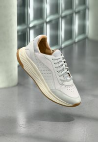 Λευκό και μπεζ sneaker BOSS με διάτρητες λεπτομέρειες, μπεζ σόλα και κορδόνια, παρουσιάζεται να αιωρείται πάνω από γκρι ανακλαστικό πάτωμα.