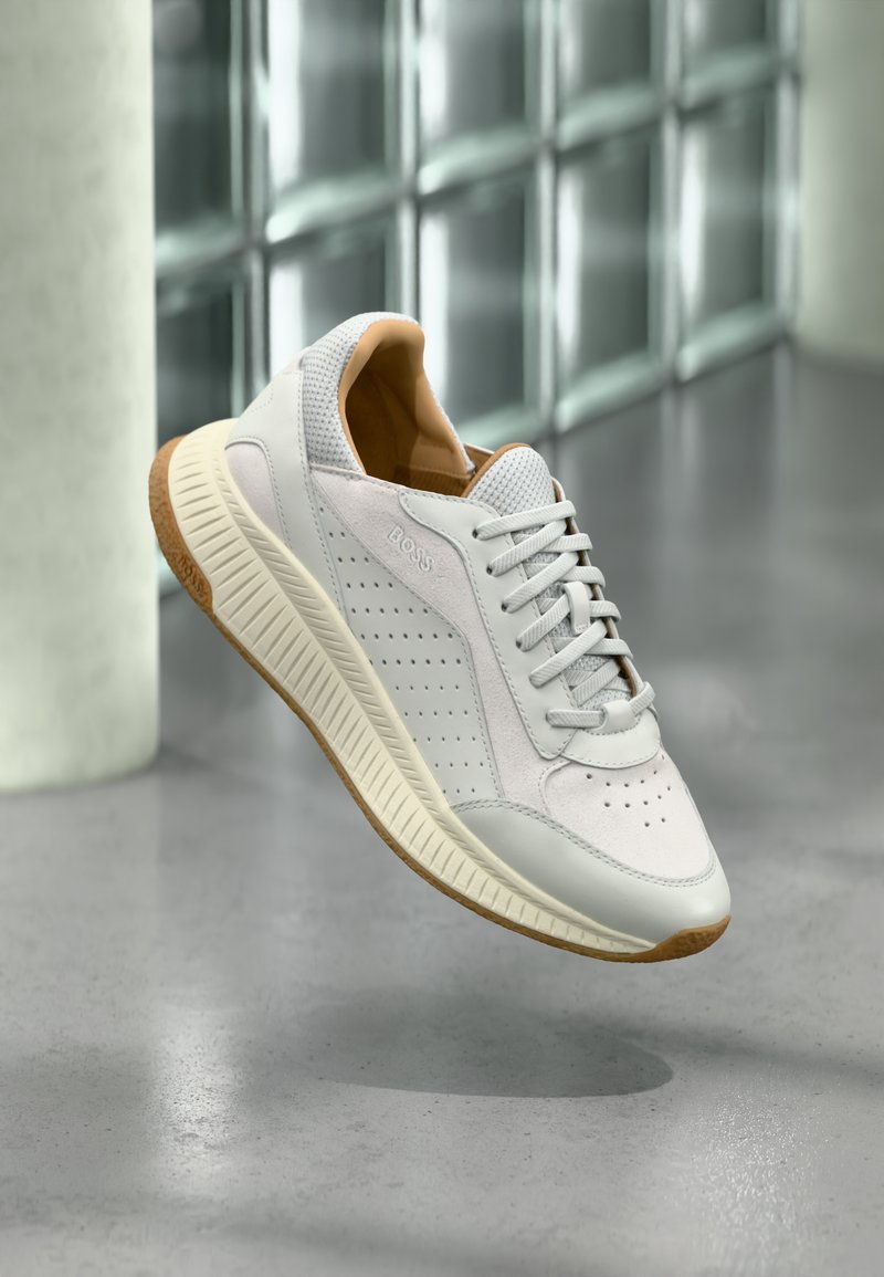 Λευκό και μπεζ sneaker BOSS με διάτρητες λεπτομέρειες, μπεζ σόλα και κορδόνια, παρουσιάζεται να αιωρείται πάνω από γκρι ανακλαστικό πάτωμα.