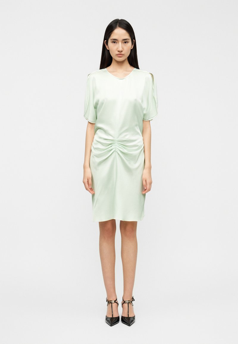 Φόρεμα από σατέν σε ανοιχτό πράσινο χρώμα με κοντά, φουσκωτά μανίκια. Διαθέτει ρυθμιζόμενη λεπτομέρεια στη μέση και καταλήγει ακριβώς πάνω από το γόνατο. Συνδυάζεται με μαύρες γόβες.