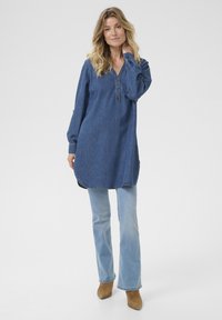 Denim tunika z V-izrezom in gumbi, z dolgimi rokavi, ohlapnim krojem in rahlo ukrivljenim robom. Oblečena s svetlo modrimi širokimi kavbojkami.