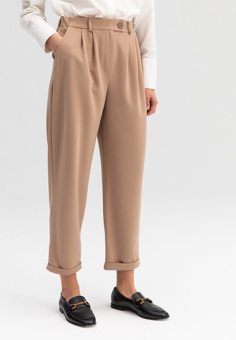 Personne portant un pantalon beige taille haute avec ourlets retroussés, une chemise blanche et des mocassins noirs avec des détails dorés, debout avec une main dans la poche.