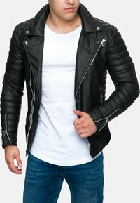 Giubbotto da moto in pelle nera con spalle imbottite, zip frontale asimmetrica e dettagli in metallo argentato. Indossato sopra una t-shirt bianca e jeans blu.