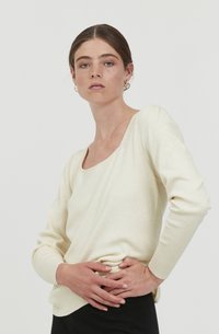 Helles Cremefarbenes Langarmoberteil mit weitem Halsausschnitt und schmalen Ärmeln. Der Stoff wirkt weich mit einer glatten Textur und feinen Rippungen entlang der Kanten.