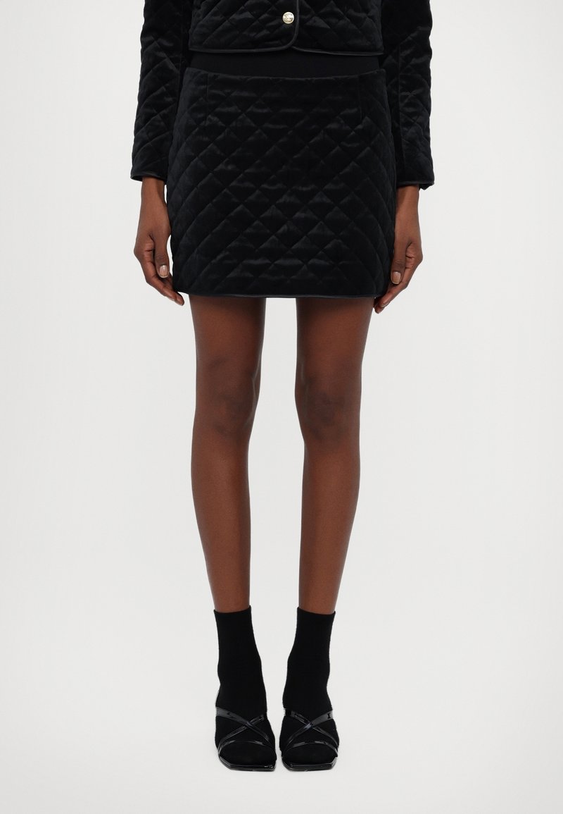 Femme portant une mini-jupe matelassée noire, une veste courte assortie, des chaussettes noires et des sandales à talons noirs à brides, debout devant un fond blanc.