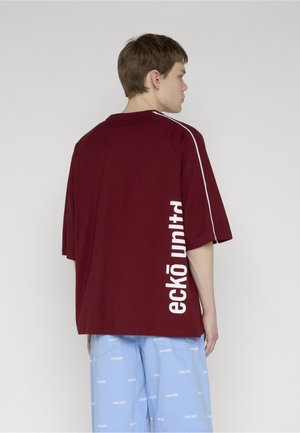 Mladý muž s krátkymi hnedými vlasmi, ktorý má na sebe voľnú vínovú košeľu s bielym nápisom „eckō unltd“ a svetlomodré šortky s rovnakým nápisom.