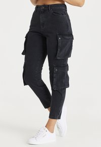 Pantalones cargo de mezclilla negra con un corte ajustado, que cuentan con múltiples bolsillos laterales, cierres a presión y un largo recortado. Combinados con zapatillas blancas.