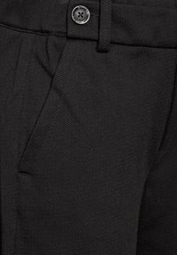 Pantaloni neri realizzati in tessuto testurizzato, con tasche laterali e chiusura a bottoni argentati. La vita presenta cuciture sottili e dettagli minimi.