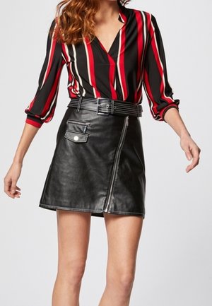 Femme portant un chemisier à manches longues rayé rouge, blanc et noir, rentré dans une mini-jupe en cuir noire avec une fermeture éclair frontale et une ceinture.