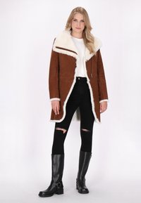 Brun ruskindjakke med en hvid pelskrave, med lynlås detaljer og en buet kant. Båret over en hvid skjorte og sorte slidte skinny jeans.