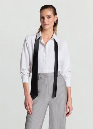 Chemise blanche boutonnée avec manches longues retroussées et une cravate en satin noir drapée autour du col ; associée à un pantalon gris à fines rayures.