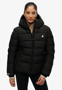 Sort pufferjakke med lynlås, der har hætte, quiltet design, sidelommer og et logo. Ribbet manchetter og strikket krave.