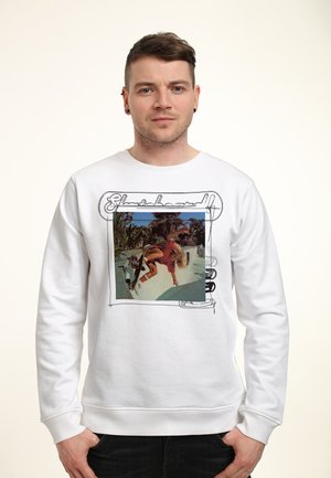 Biała bluzka z nadrukiem graficznym przedstawiającym deskorolkarza otoczonego palmami, z czarnym kursywą napisem "Skateboard."