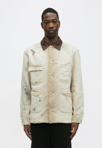 Veste en toile beige avec un col marron, deux poches avant, des détails usés et des accessoires en métal argenté. Texture douce avec des marques dispersées.