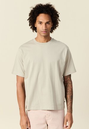 Mężczyzna z kręconymi włosami, ubrany w gładką beżową koszulkę i jasnoróżowe spodnie, stojący na beżowym tle, z tatuażami na prawym ramieniu.