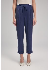 Pantaloni blu navy con vita regolabile, pieghe e orli arrotolati. Realizzati in un tessuto leggero. Abbinati a scarpe con tacco color crema.