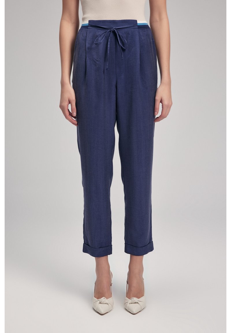 Pantaloni blu navy con vita regolabile, pieghe e orli arrotolati. Realizzati in un tessuto leggero. Abbinati a scarpe con tacco color crema.