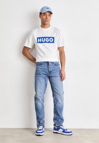 Tricou alb cu logo-ul albastru "HUGO", șapcă albastră deschisă, blugi albaștri deschiși și adidași albi cu accente albastre. Fundal simplu.