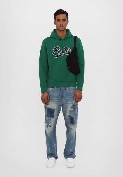 Felpa verde con cappuccio e logo bianco, jeans azzurri chiari strappati con macchie scure, e sneakers bianche. Borsa a tracolla nera.