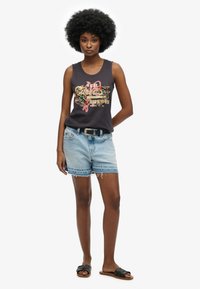 Débardeur graphique sans manches noir avec un design coloré, associé à un short en denim bleu clair avec un ourlet effiloché et des coutures contrastées. Sandales noires à glisser.