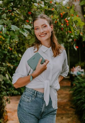 Green zippered wallet with a smooth texture, held in a hand. White button-up shirt styled with a knotted front and light blue high-waisted jeans.