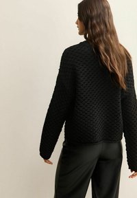 Zwarte gebreide trui met een textuurpatroon en lange mouwen, gecombineerd met elegante zwarte broeken. De achterkant toont een cropped ontwerp.