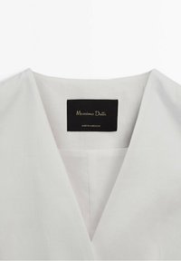 Biała bluzka z dekoltem w kształcie litery V, o fakturowanej tkaninie, z czarną metką z napisem "Massimo Dutti, wyprodukowane w Maroku" przy dekolcie.