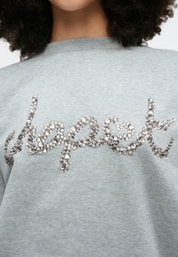 Pelēks džemperis ar fakturētu, metālisku sudraba uzrakstu "cerība", rotātu ar dažāda izmēra strass akmentiņiem. Mīksts audums, brīvs piegriezums.