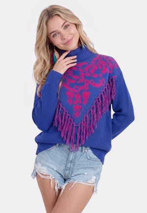 Lyshåret kvinde iført blåt strik med pink blomstermønster og frynser, kombineret med slidte lyse denimshorts, smilende med hånden nær ansigtet.
