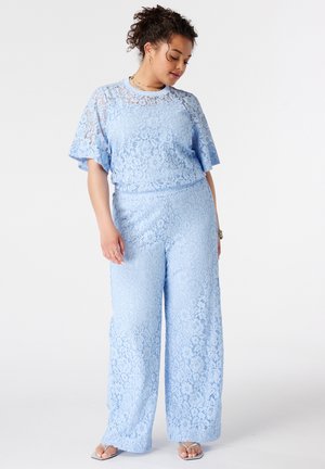 Femme portant un haut en dentelle bleu clair à manches courtes et un pantalon large avec des sandales, debout sur un fond blanc uni.