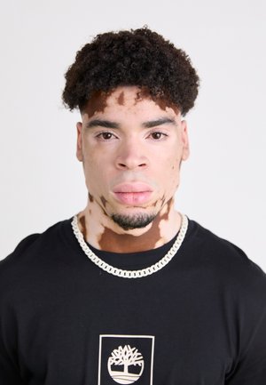 Svart T-skjorte med grafisk logo, sølvkjede, kort krøllete hår og variert hudfarge med flekker av lyse og mørke toner.