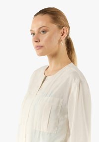 Jasnoszara bluzka z długim rękawem, zamek błyskawiczny i dwoma kieszeniami na piersi. Gładka tekstura i luźny krój. Model pokazany w profilu.