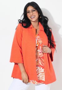 Světlý oranžový strukturovaný kabát s límcem a zapínáním na knoflíky. Nosit na květinovém vzoru topu s růžovými a oranžovými tóny. Bílé kalhoty.