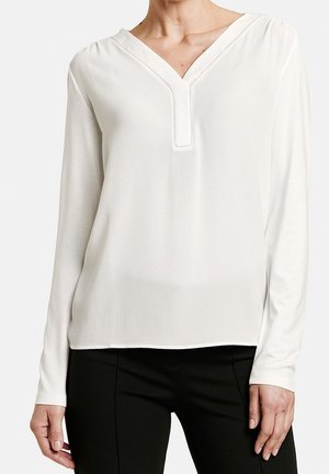 Femme portant un chemisier blanc à manches longues avec un col en V, associé à un pantalon noir, visible de la nuque jusqu'aux hanches sur un fond uni.