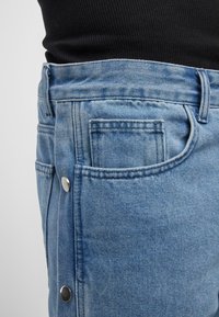 Helleblaue Denim-Jeans mit seitlichen Knöpfen, ausgestattet mit zwei Vordertaschen und einem strukturierten Stoff. Der Bund ist leicht erhöht.