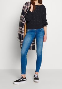 Suéter de punto de cable azul marino, jeans ajustados azules y zapatillas negras con suelas blancas. El modelo sostiene un abrigo de patrón de cuadros.