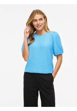 Femme souriante, portant un haut bleu clair texturé à manches courtes bouffantes et un pantalon noir, main près des cheveux, debout devant un fond blanc.