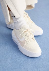 Bílé kožené tenisky Nike se světle béžovými akcenty, plochými tkaničkami a texturovanou podrážkou. Na nohou jsou prezentovány s ribkovanými bílými ponožkami a béžovými kalhotami.