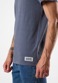 Camicia a maniche corte grigia con trama intrecciata, con etichetta sull'orlo, abbinata a jeans di denim blu.