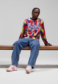 Röd- och gulfärgad tie-dye långärmad tröja med vit logotyp, kombinerad med blå jeans och vita sneakers med röda ränder, sittande på en bänk.