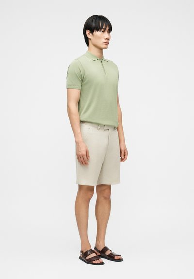 Άνδρας που στέκεται φορώντας ανοιχτό πράσινο κοντομάνικο πόλο, μπεζ σορτς και σκούρα καφέ σανδάλια, μπροστά σε απλό λευκό φόντο.