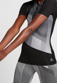 Svart kortärmad träningströja med diagonala grå ränder och textilstruktur; tillsammans med svart och vit randiga leggings med Reebok-logotyp.