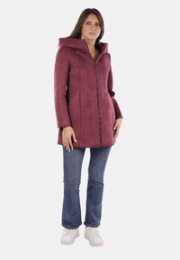 Cappotto borgogna con cappuccio e zip davanti, texture morbida e maniche lunghe, abbinato a jeans blu e sneakers bianche.