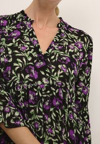 Robe noire avec un motif floral violet et vert, dotée d'un décolleté en V et d'une bordure froncée, fabriquée dans un tissu texturé avec des manches évasées.