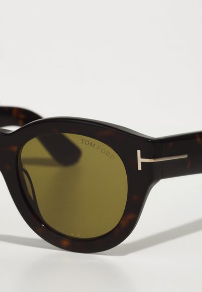 Ochelari de soare cu ramă din scoică țestoasă, cu lentile verzi și elemente din metal argintiu. Au inscripția "TOM FORD" pe lentila stângă.