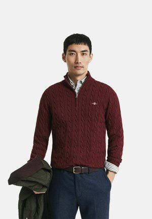 Burgunderfarbener Kabelstrickpullover mit einem Viertelreisverschluss, über einem Hemd mit Kragen getragen. Das Modell hält eine grüne Jacke und steht vor einem weißen Hintergrund.