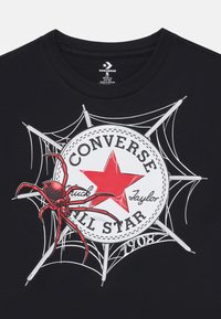 Un t-shirt noir présentant un graphique d'araignée et de toile, mettant en valeur un logo Converse en cercle avec une étoile rouge et du texte.