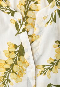 Blouse blanche avec un motif floral jaune comprenant des tiges vertes, un petit col et un devant avec un col en V boutonné.