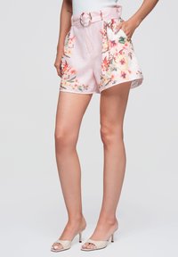 Femme portant un short taille haute à fleurs rose clair avec une ceinture, associé à des sandales beiges à talons ouverts, debout devant un fond gris.
