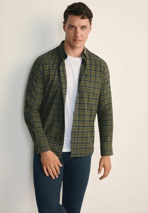 Zielona koszula w kratę z flaneli, z kołnierzykiem zapinanym na guziki, długimi rękawami i kontrastowymi niebieskimi liniami, noszona na białej koszulce oraz ciemnych spodniach.